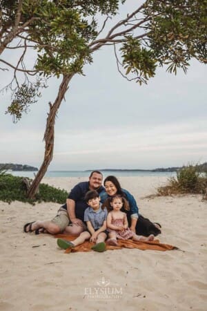 A family sit under a tree on a blanket on a sandy beach