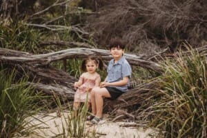 Two children sit together on a log surrounded by trees on a beach