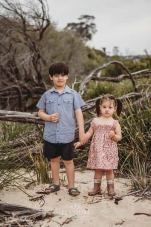 Two siblings stand holding hands on a beach with trees behind them
