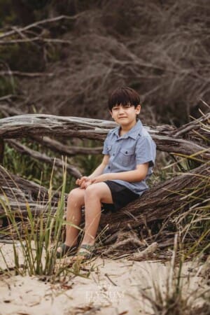 A boy sits on a log at a beach with driftwood and bushes behind him