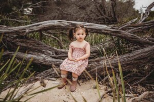 A little girl sits on a large driftwood log at a beach surrounded by bushes