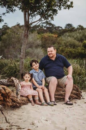 A father sits on a fallen tree with his kids on a sandy beach