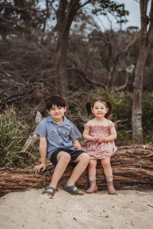 Two kids sit on a large fallen tree on a sandy beach at sunset