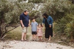 A family hold hands with their children as they stand on a beach surrounded by trees