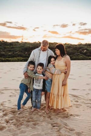A family stand cuddling their three childnen on a Cronulla sand dune at sunset