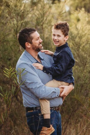 A dad holds his son as they laugh together in a bushy field at sunset