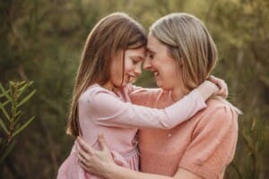 A mother hugs her daughter and they gently press their faces together smiling with the sun setting behind them