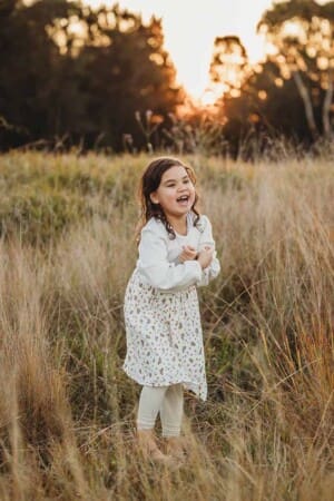 A warm photograph of a little girl laughing as she stands amongst long grass at sunset