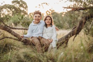 Two children sit on a low tree branck that stretches over long grass with the sun setting behind them