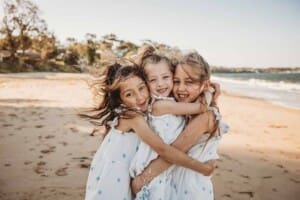 Three little girls with windswept hair stand hugging on a sunny beach