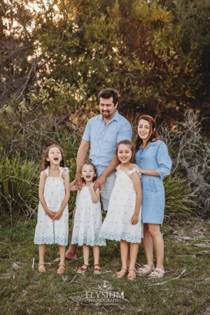 A family of five stand hugging on a beach surrounded by trees