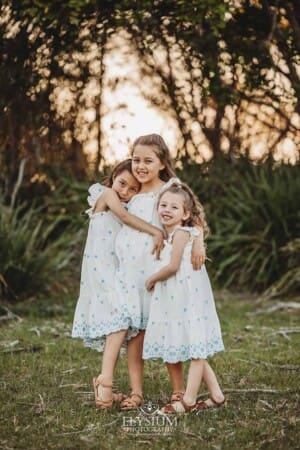 Three little girls stand on a beach hugging surrounded by trees at sunset