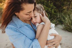 A little girl is cuddled by her mother as they sit on a beach at sunset