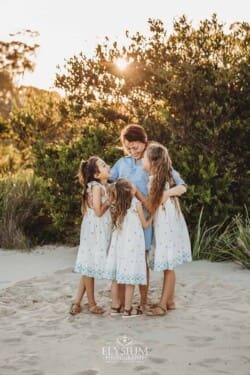 A mother hugs her three daughters as they stand on a tree lined beach at sunset