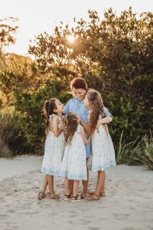 A mother stands on a beach hugging her three daughters as the sun sets behind them