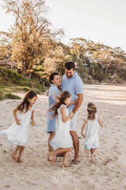 A couple stand on a beach hugging as their three girls run in circles around them