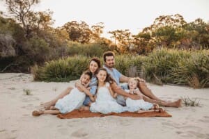 A family sit on a blanket on a beach and hug each other at sunset