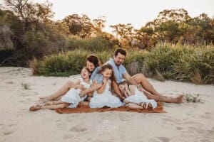 Parents tickle their three girls as they sit on a blanket on a beach at sunset