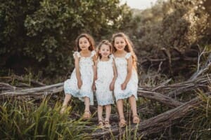 Three siblings sit on a fallen tree branch surrounded by bushes at sunset