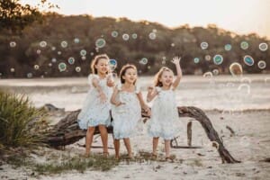 Three little girls sit on a fallen tree and blow bubbles at sunset on a beach