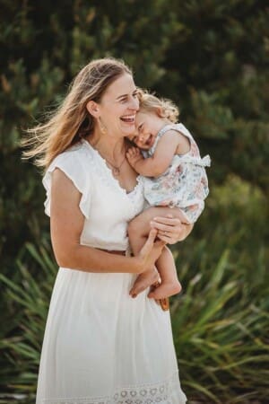 A mother cuddles her giggling baby girl as they stand in a bushy area at sunset
