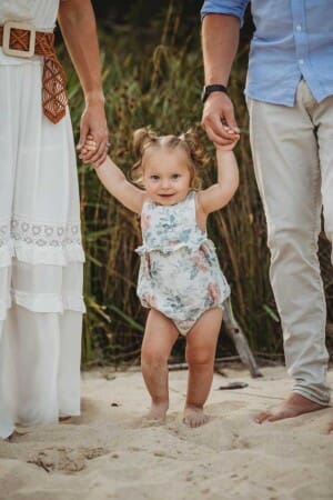 A baby girl stands on the sand between her parents holding their hands and smiling