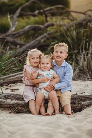 Three children sit on a log at a beach and smile as the sun sets behind them