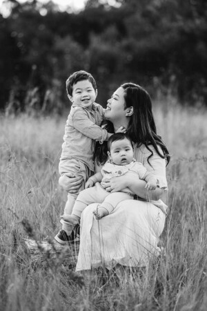 A mother sits on a log in a grassy field with her baby boys in her lap enjoying the sunset