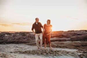 A pregnant woman and her husband walk together along a beach at sunset for a maternity session in Cronulla