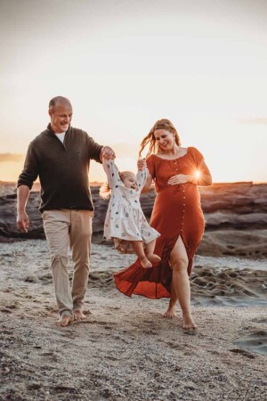 Parents lift their smiling baby girl between them as they walk along a beach at sunset during a family session at Cronulla