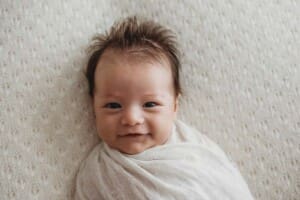 A baby boy with spikey hair smiles as he lays in a white wrap on a matching blanket