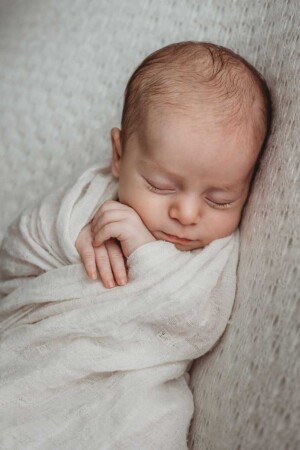 A wrapped newborn baby lays sleeping on a textured white backdrop