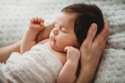 A mother's hands hold her sleeping newborn baby laying wrapped on a textured white blanket