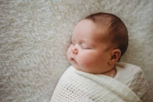 A newborn girl lays sleeping in a soft white wrap on a matching white blanket