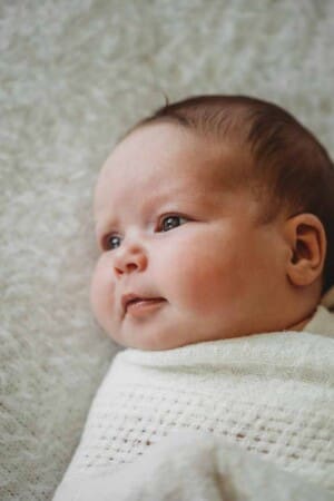 A baby girl smiles softly as she lays on a white blanket wrapped in a matching white cloth