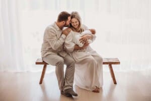 Parents sit on a wooden bench cradling their newborn baby between them in front of a bright window