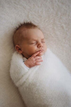 A newborn baby lays sleeping wrapped in a white blanket with it's finger pressed against its mouth