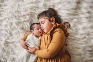 A little girl lays on a white fur rug cuddling her newborn baby sister
