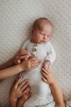 Siblings place their hands on their newborn baby sister as she lays sleeping on a white blanket