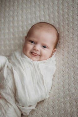 A newborn baby pokes her tongue out with a cheeky little grin laying on a white blanket