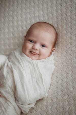 A newborn baby pokes her tongue out with a cheeky little grin laying on a white blanket