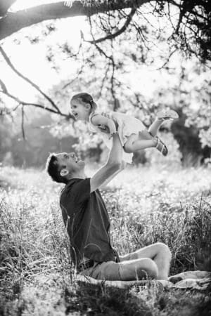 A dad sits on a blanket in a grassy field and lifts his daughter above his head as she giggles