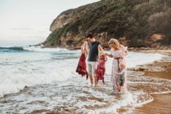 A family of five stand in the water at a beach and hold their childrens hands as they all have fun jumping the waves