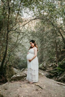 A pregnant woman holds her belly as she stands on a large rock surrounded by trees