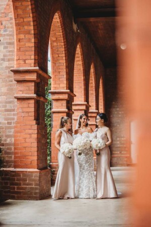A bride and bridesmaids stand amongst brick arches at a church before the wedding ceremony
