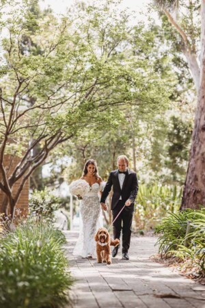 A bride and groom walk their dog through a lush green garden after their wedding ceremony