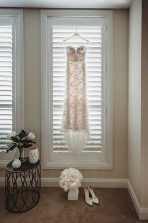 A brides dress hangs in a window with her shoes and bouquet sitting on the floor