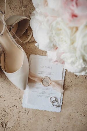 A brides shoes, bouquet, rings and wedding invitation sit on a stone floor