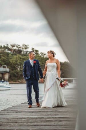 A bride and groom walk holding hands along a pier as the sun sets behind them
