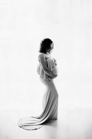 A maternity image of a mother in a flowing gown stands in front of a large window holding her pregnant belly